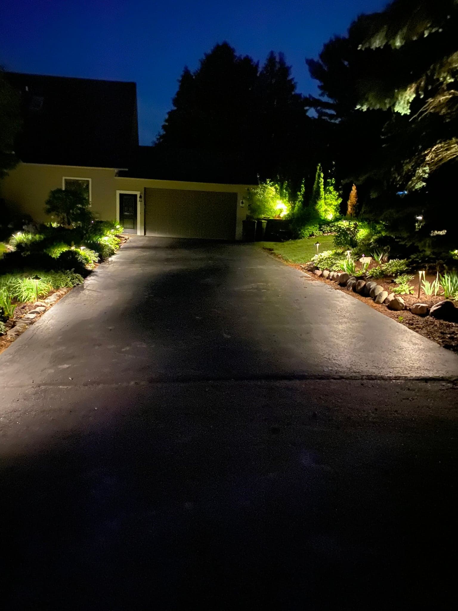 Residential driveway at night flanked by uplit landscape plantings, path lights, and warm-toned beds — landscape lighting installation