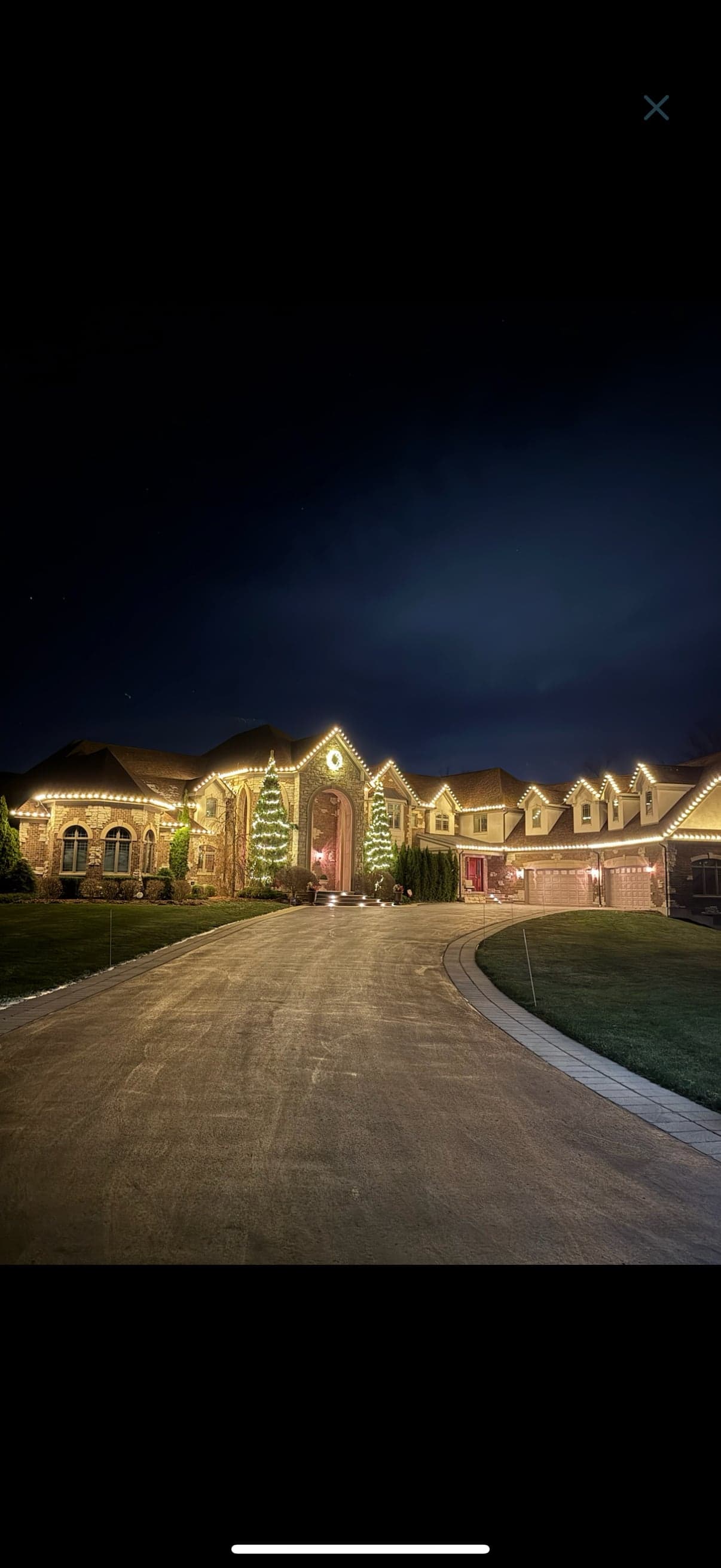 Large stone-and-brick estate with extensive warm-white holiday lighting outlining every roofline, lit specimen trees flanking the entry, and an illuminated paver driveway approach