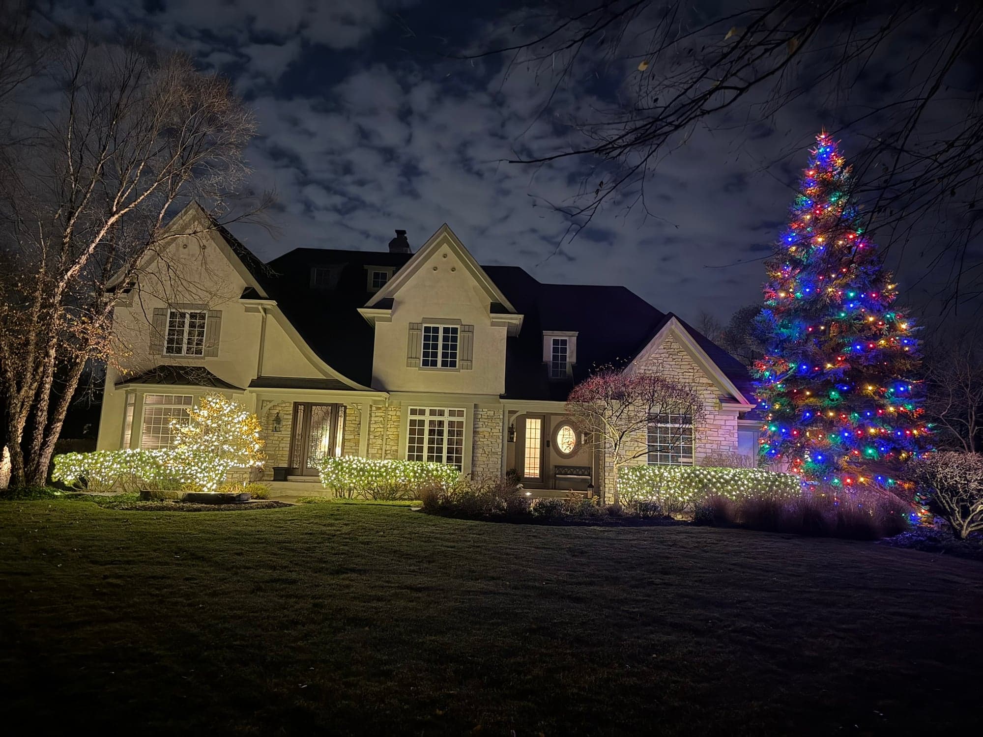 Stuccoed two-story home with a tall multicolor-lit Christmas tree, white-net-lit bushes along the front, and architectural uplighting on the stone facade