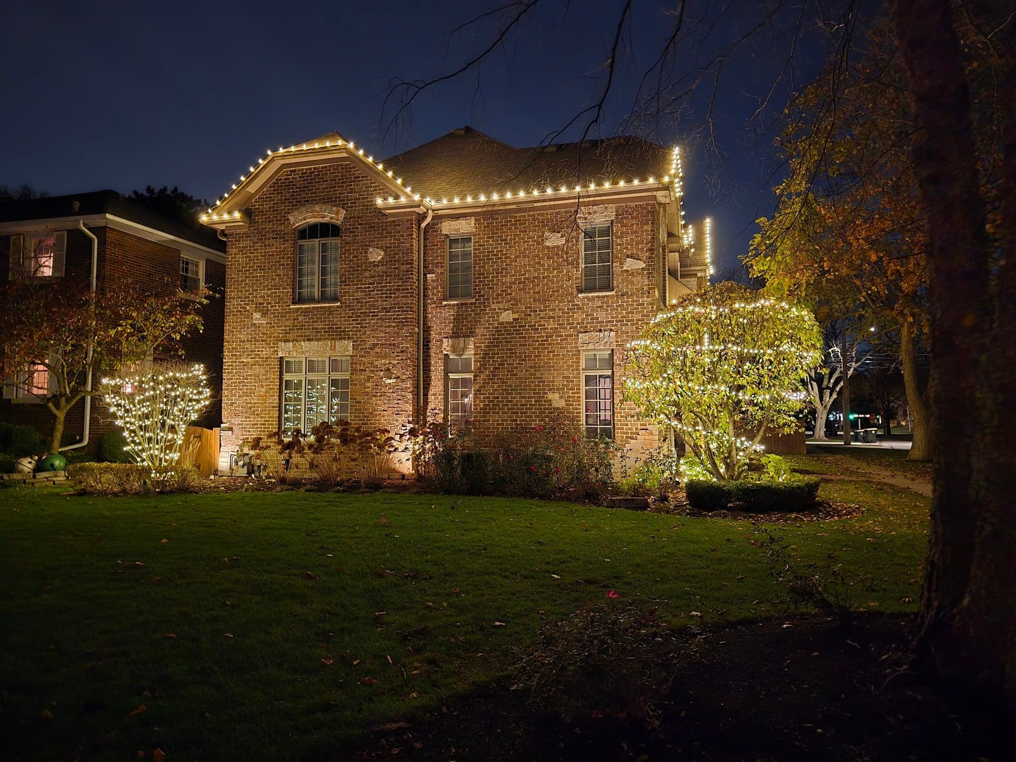 Brick home with warm-white roofline holiday lighting, illuminated front shrub, and uplighting on the facade