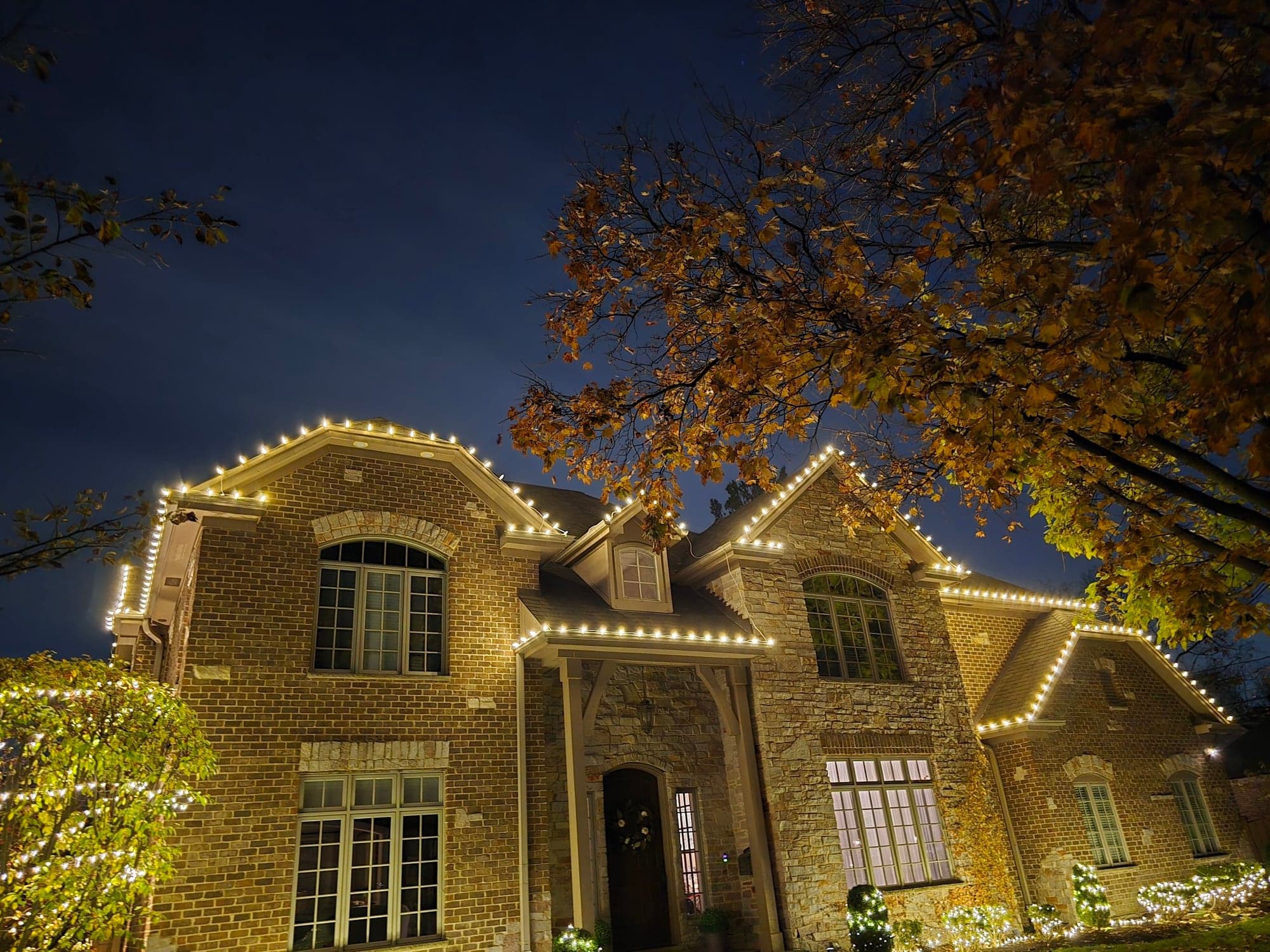 Brick two-story home with classic warm-white roofline holiday lighting, stone window arches, and lit shrubbery at the foundation