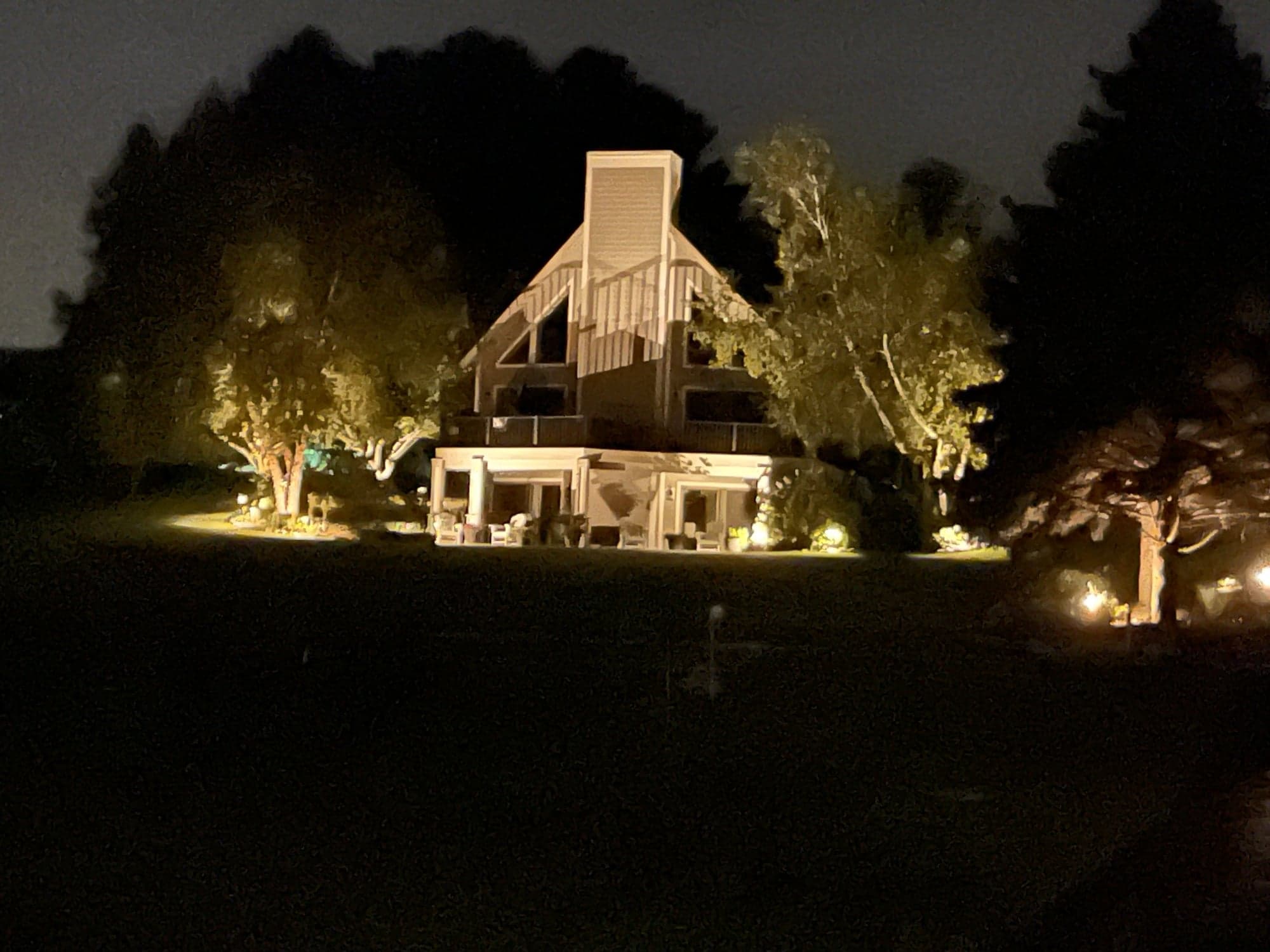 Cedar-and-stone lake home at night with architectural uplighting on the chimney and gables, and uplit specimen trees in the surrounding lawn