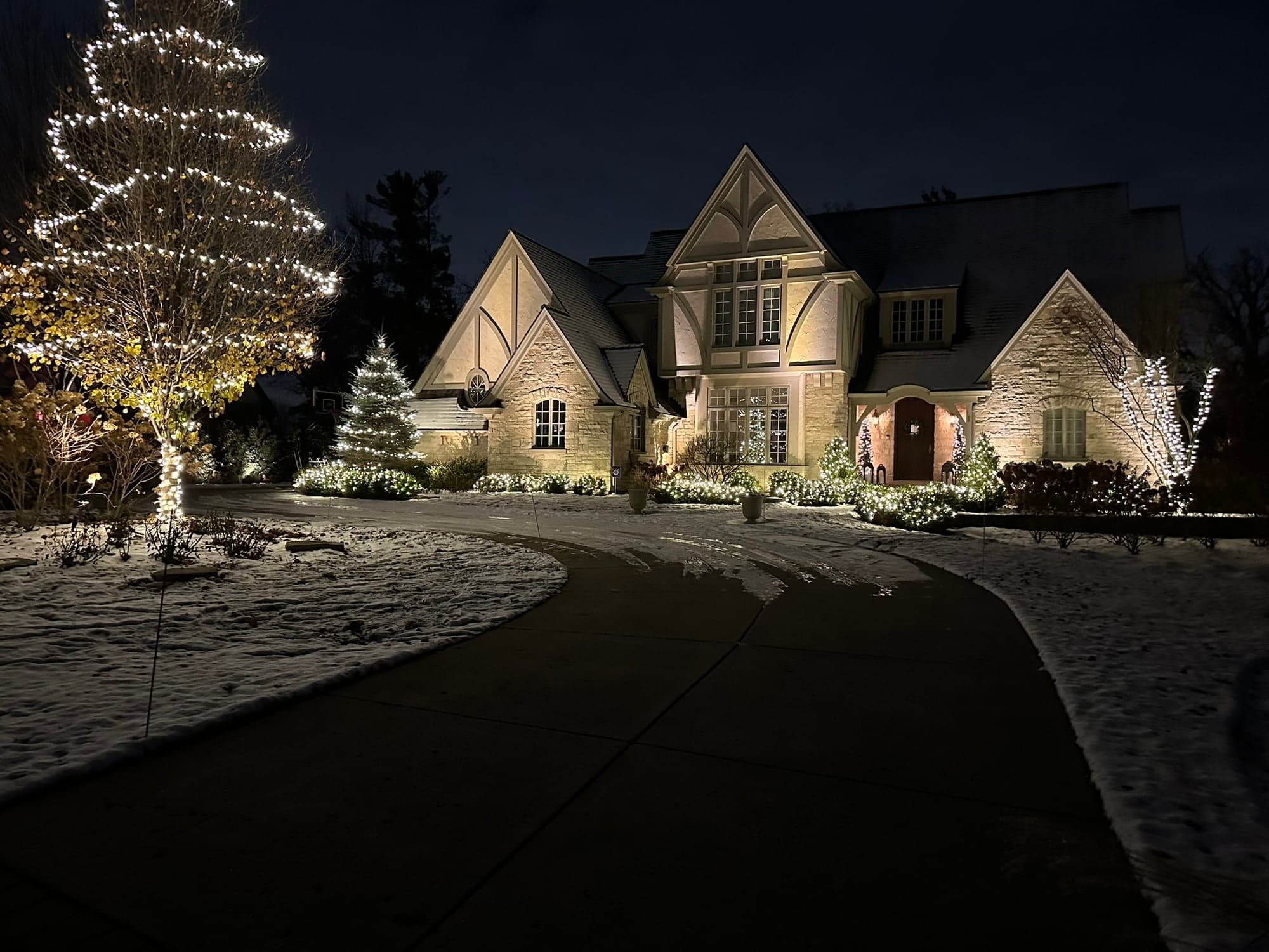 Tudor-style estate with traditional warm-white holiday lighting on the rooflines, an illuminated Christmas tree, and architectural uplighting on the stone facade, with fresh snow on the ground