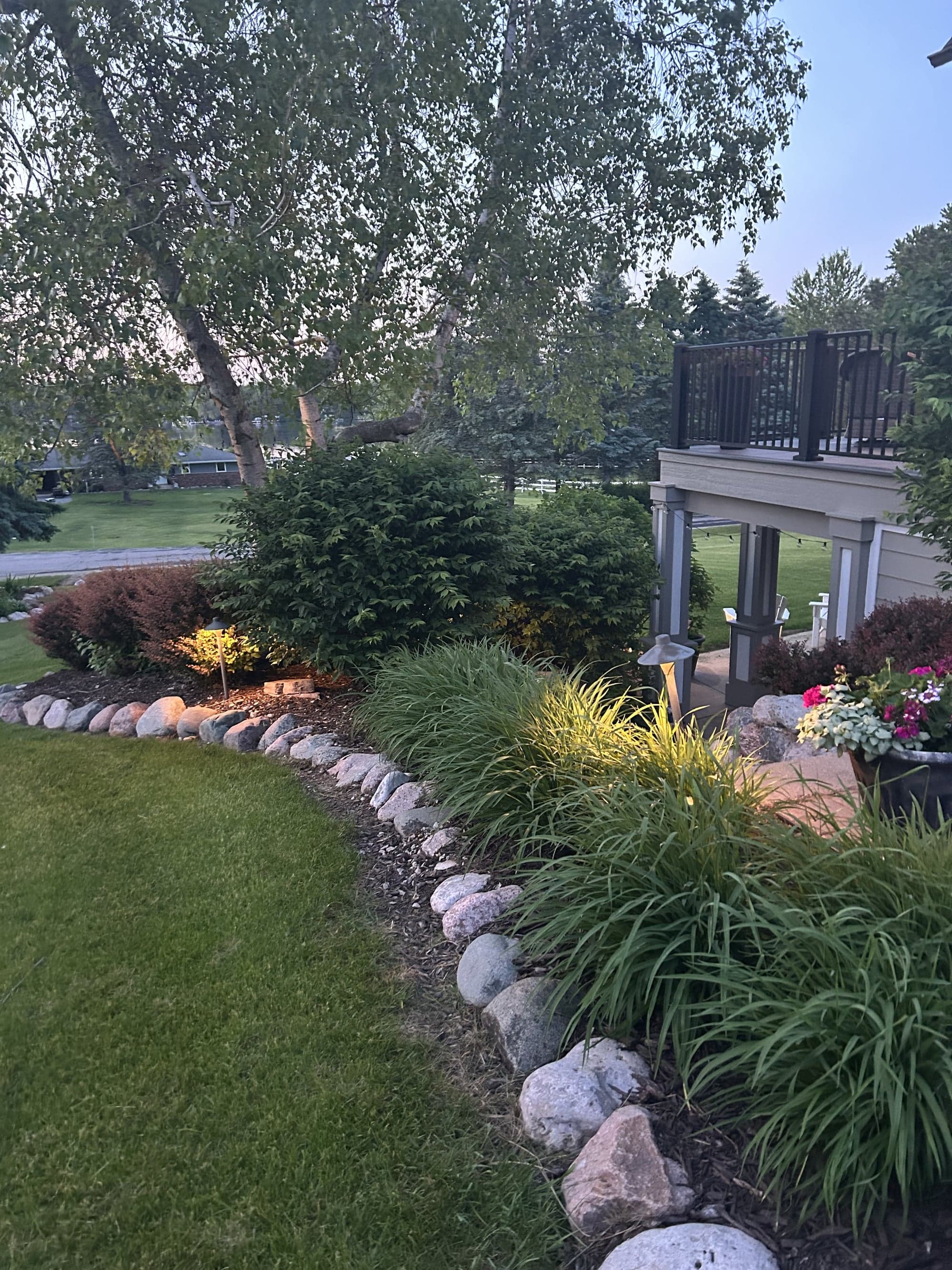 Stone-bordered planting bed with mature shrubs, uplit by warm low-voltage lights, adjoining a paver patio with seasonal container plantings