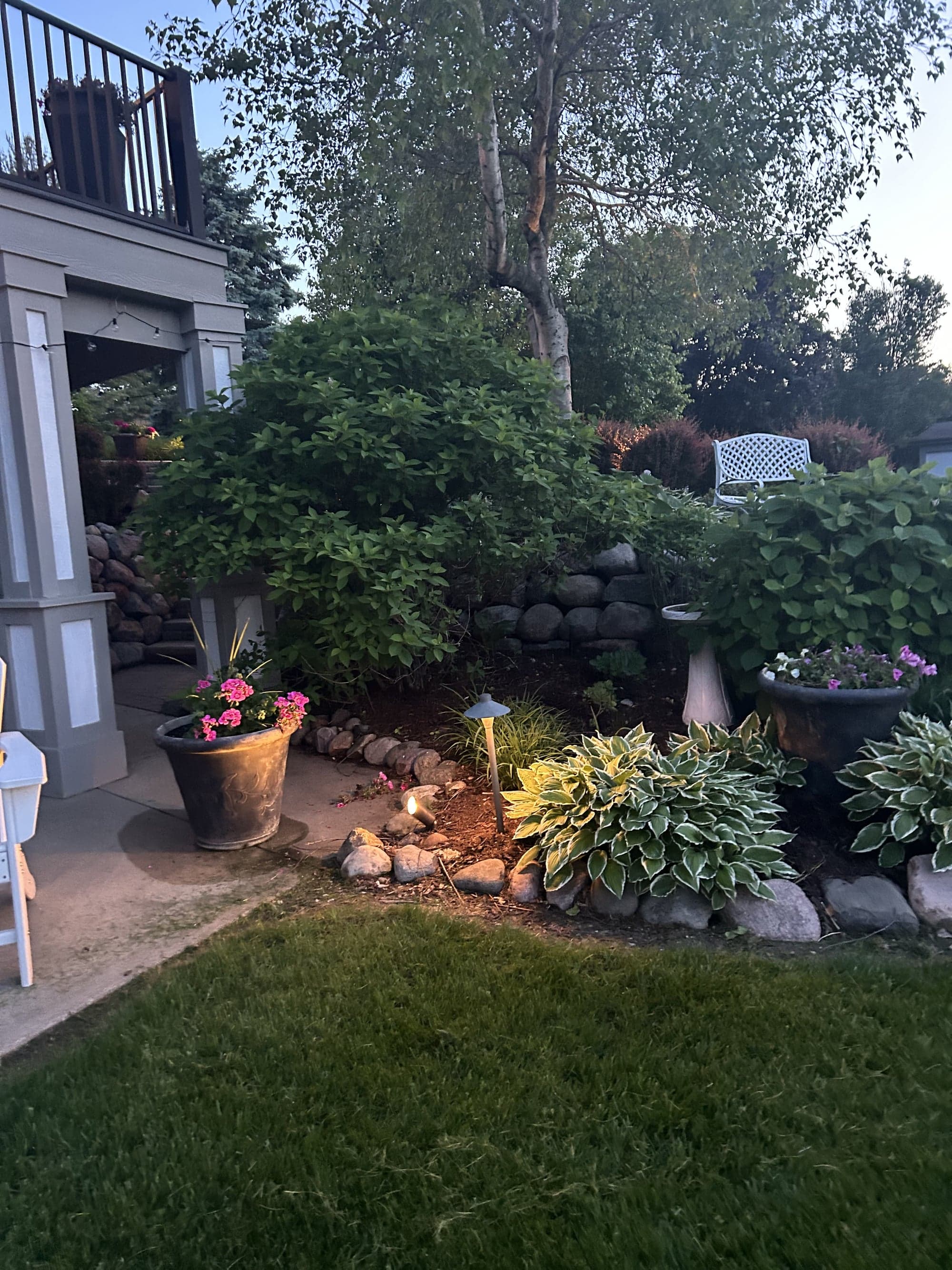 Stone-bordered planting bed beside a paver walkway, with a path light, container plantings, and a backdrop of mature shrubs