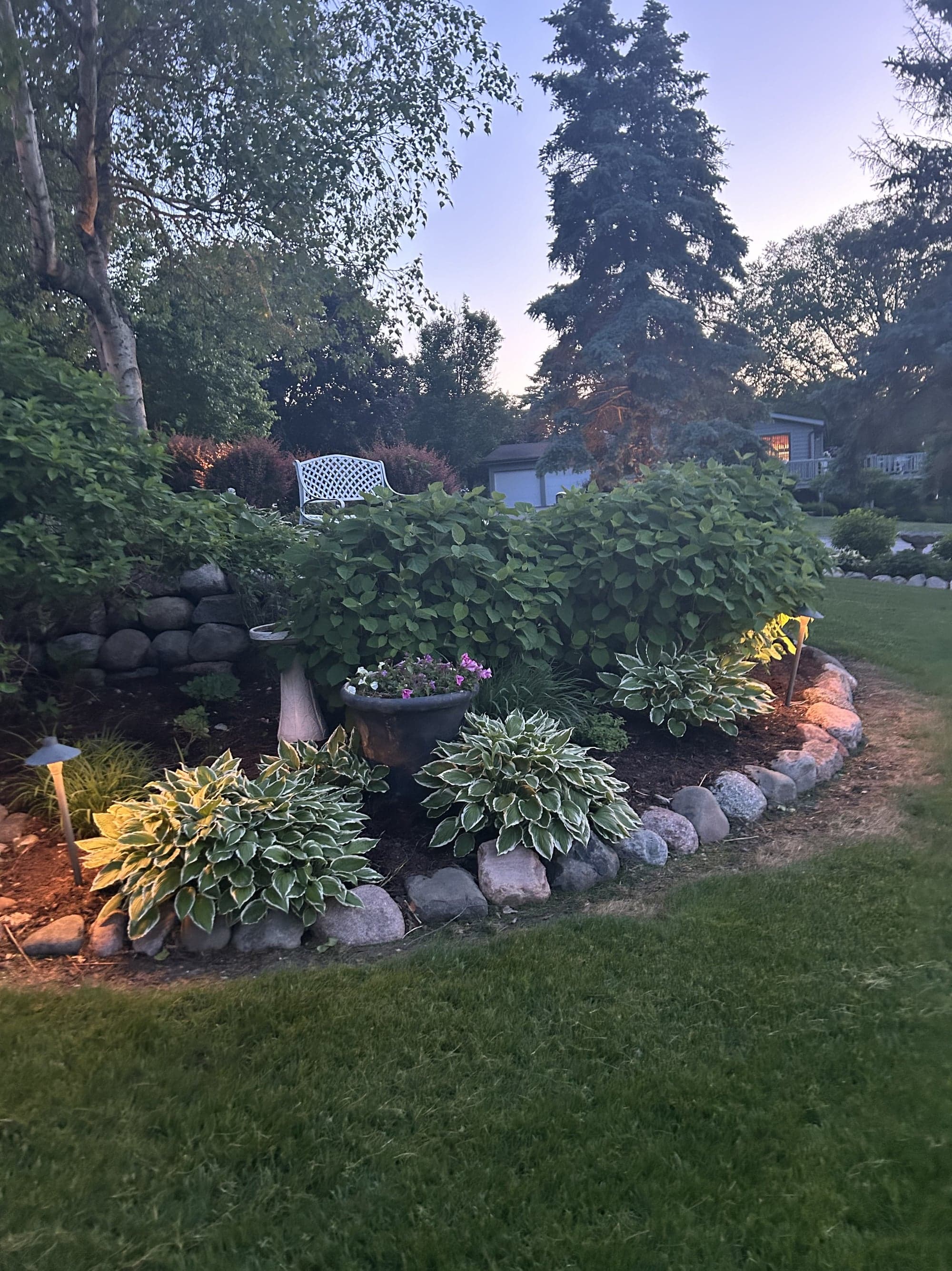 Stone-walled raised planting bed with variegated hostas and seasonal flowers, with a path light illuminating the path edge